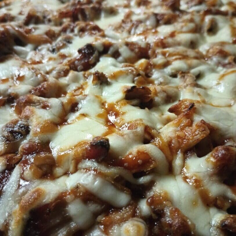 20230111_165243910_iOS A close-up of a pizza topped with melted cheese and pieces of cooked meat, reminiscent of a BBQ catering service feast, on a wooden table.