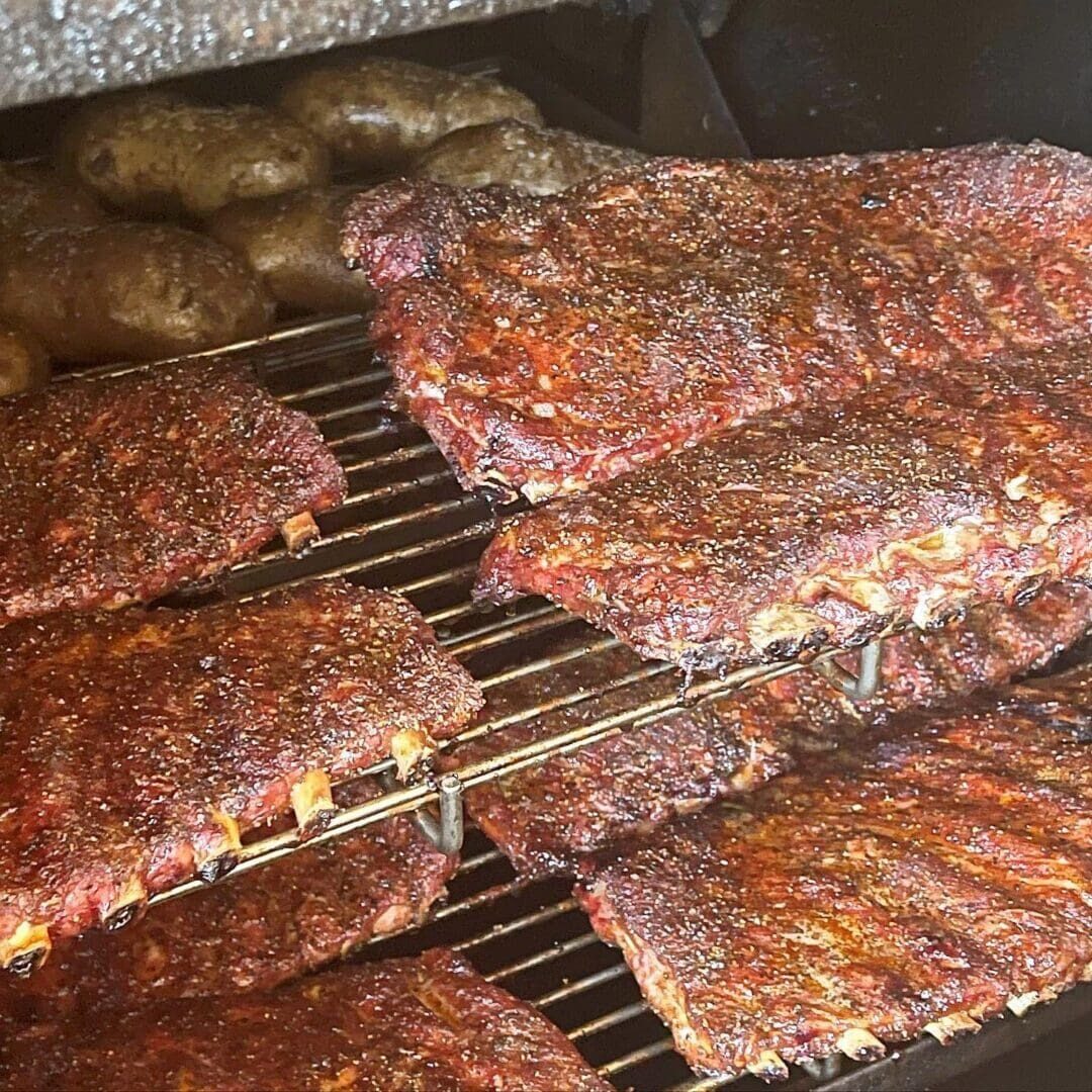 20230505_134848922_iOS Racks of seasoned ribs are being smoked on a grill by our expert BBQ catering service, with several baked potatoes positioned on an upper rack.