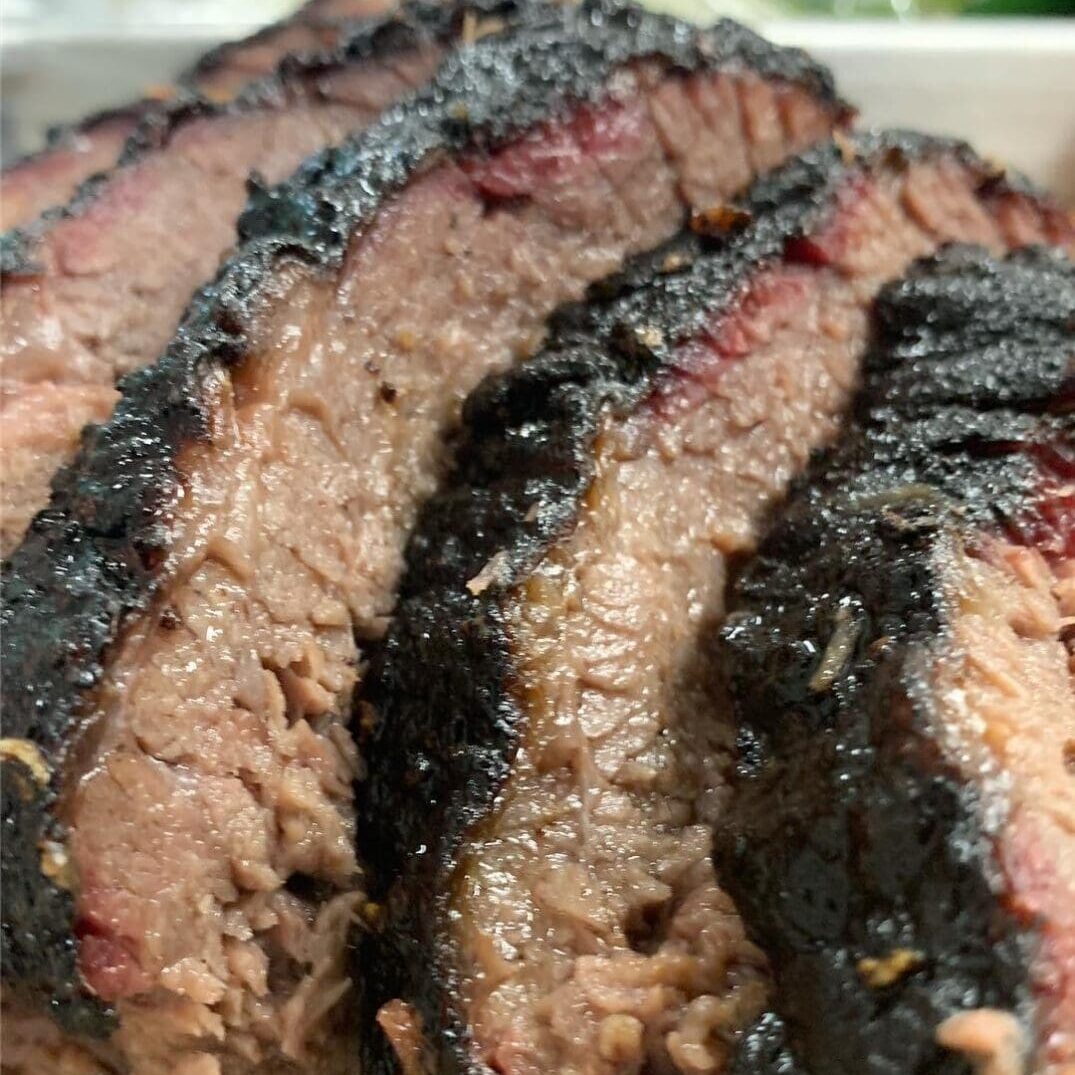 IMG_0033 Close-up of several slices of smoked brisket with a dark crust and juicy interior, expertly prepared for a BBQ catering service, stacked enticingly on a plate.